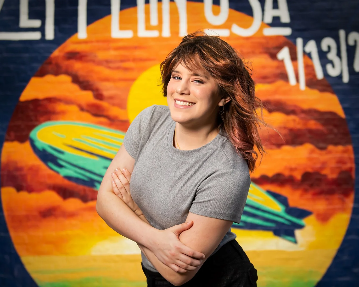A woman with short, wavy red hair, wearing a gray t-shirt, smiling with arms crossed. Behind her is a colorful mural of a sunset over the ocean in Downtown Tucson, Az