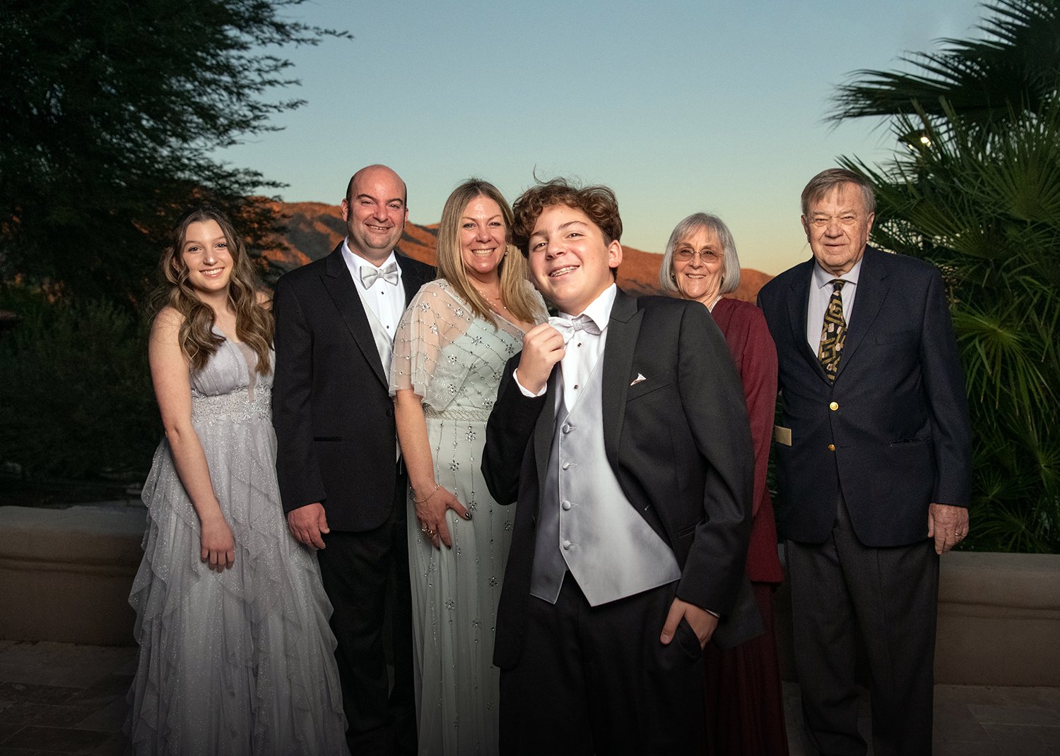 Group of six people dressed formally, standing outdoors during sunset, with hills and trees in the background.