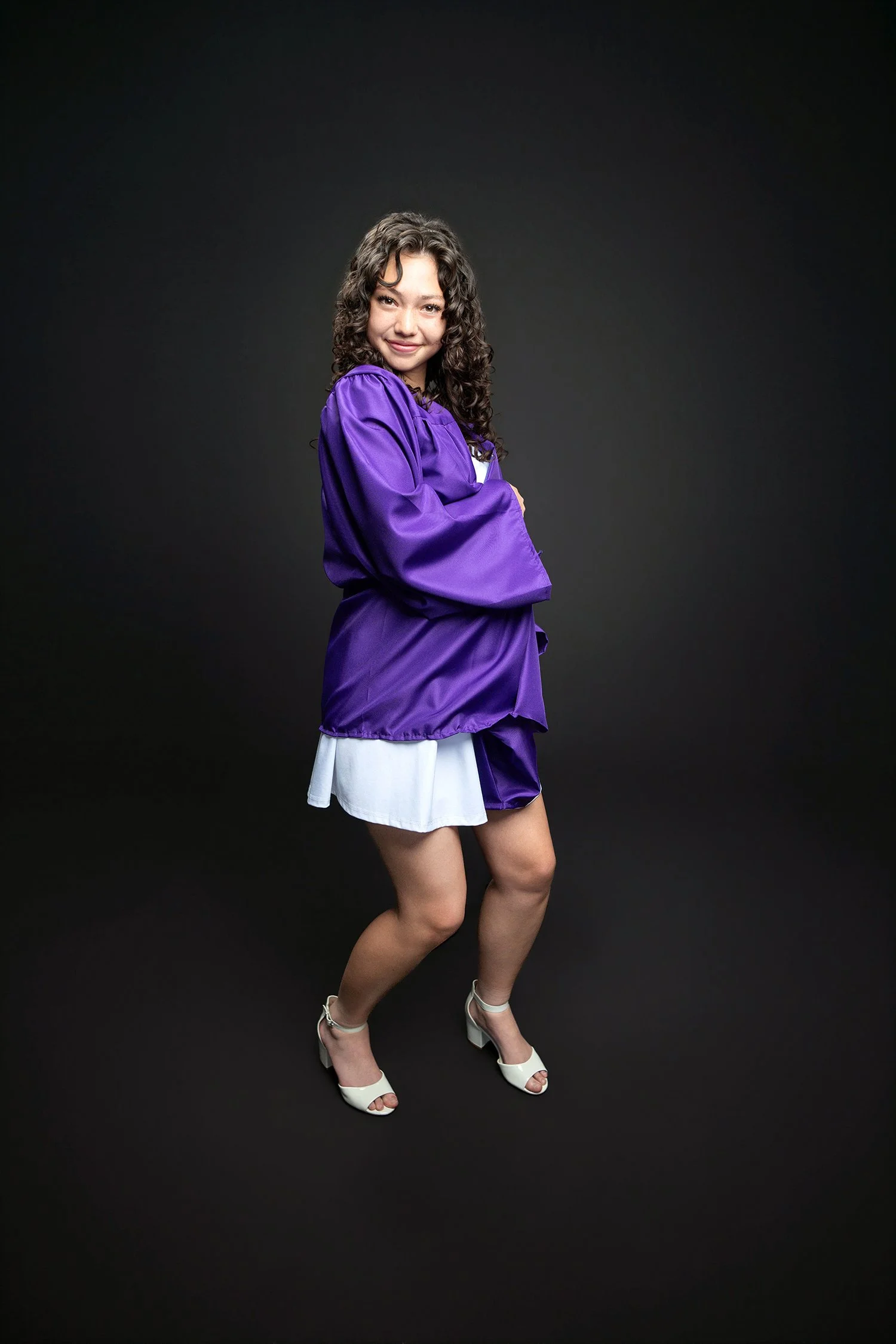 A young woman with curly hair, smiling, wearing a purple satin jacket, white top, white skirt, and white high-heeled sandals, standing against a black background.