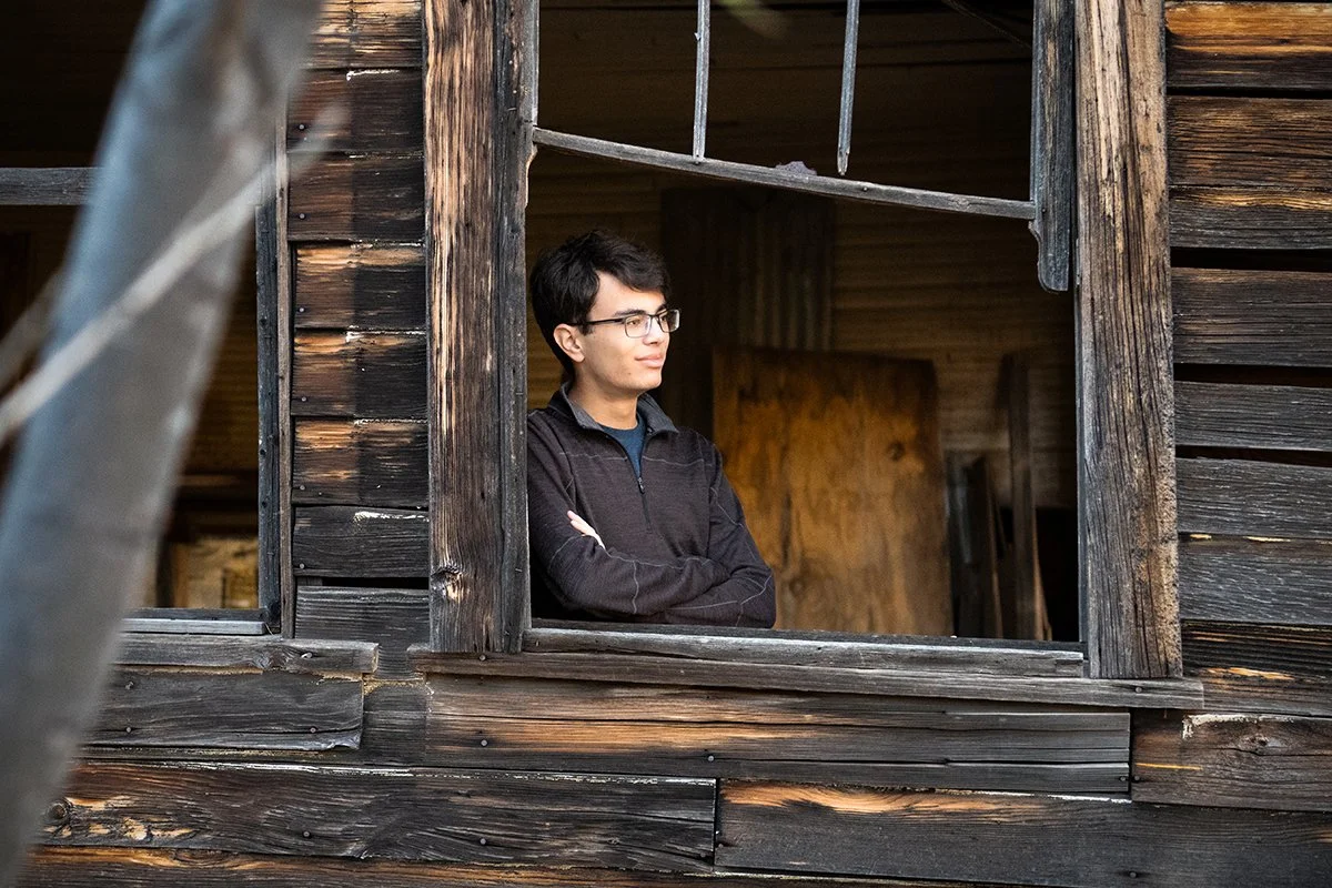 Young man with glasses leaning on a window frame of a weathered wooden building, looking outside thoughtfully during a Southern Arizona senior portrait shoot