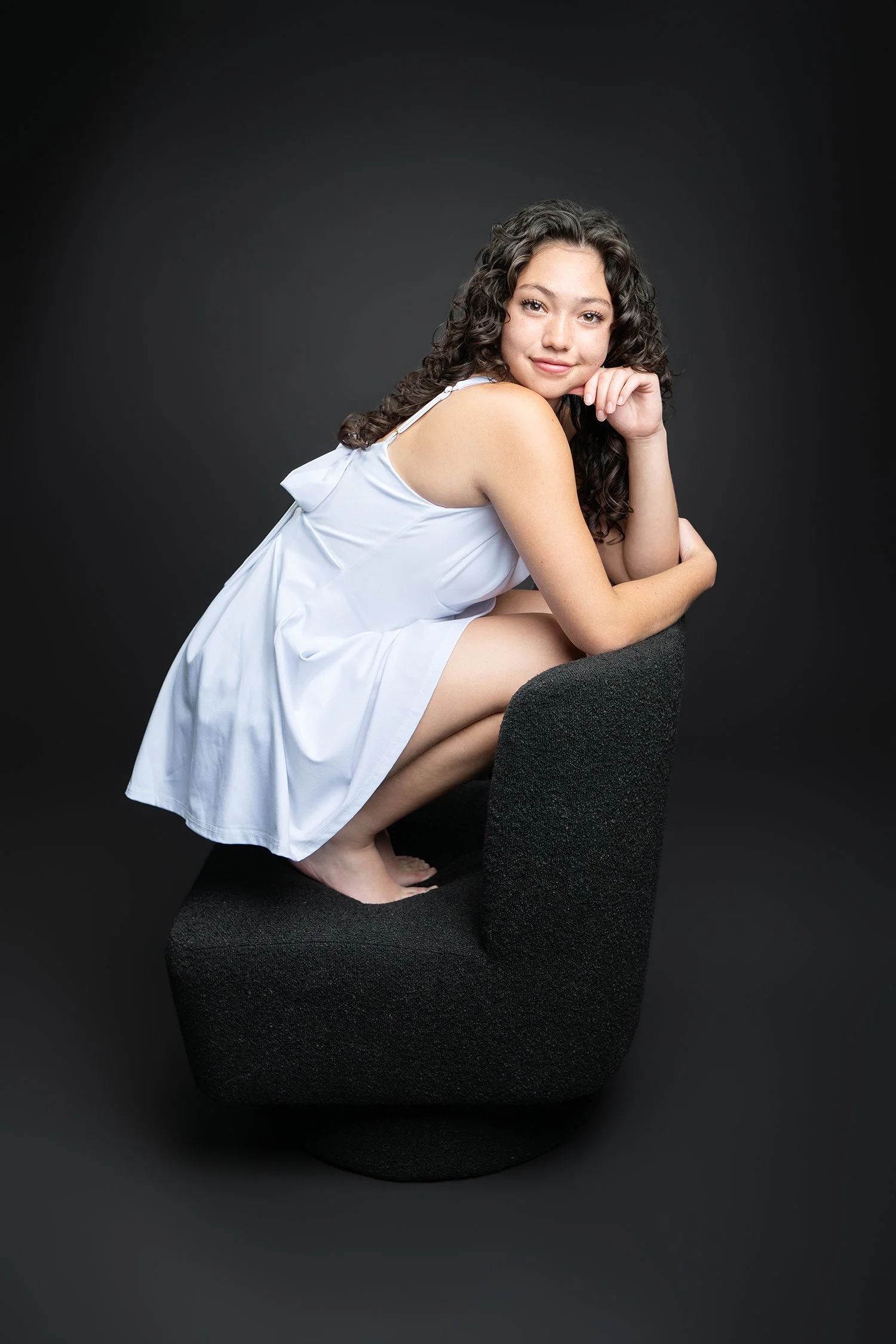 A young woman with curly hair, dressed in a white dress, sitting on a black chair against a dark background, smiling gently at the camera.
