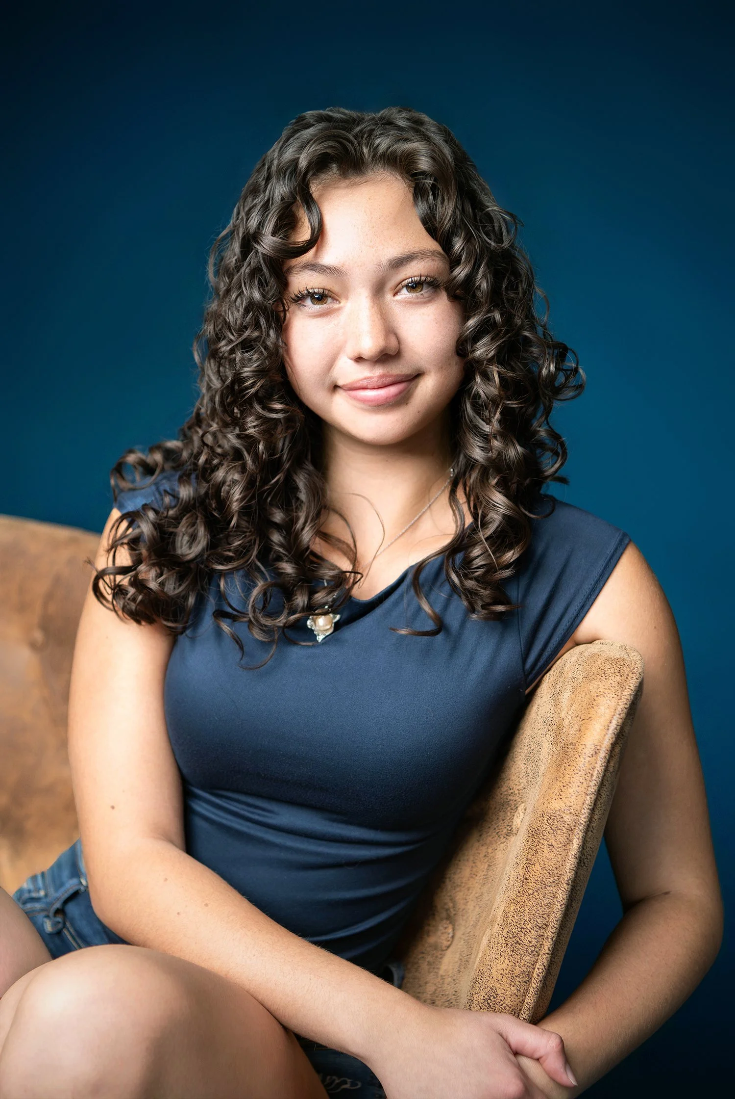 A young woman with long, curly brown hair, wearing a sleeveless navy blue top and shorts, sitting on a beige chair against a dark blue background, smiling softly at the camera.