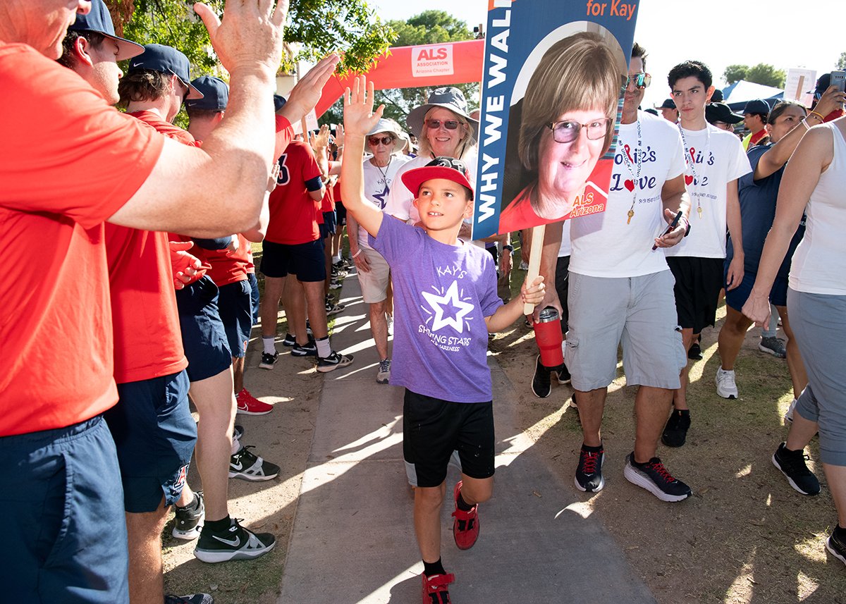A young boy holding a sign with a woman's face at a crowd rally or march. The boy wears a purple shirt, black shorts, and red shoes, and is giving a high-five to a person in the crowd. The crowd is wearing casual clothing, some with t-shirts supporti