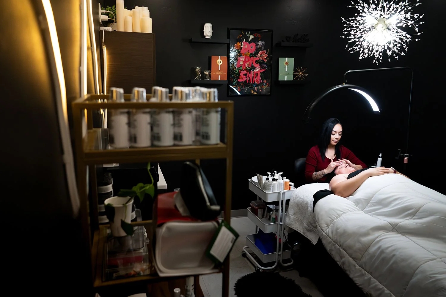 A woman receives a facial treatment in a dark, modern spa room with black walls and contemporary decor. She is lying on a bed while a technician applies a skincare product. The room has a stylish chandelier, framed artwork, and a tall lamp above the 