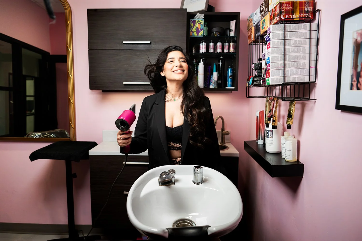 A woman with long dark hair smiling in a salon, holding a pink blow dryer, with various hair products on shelves and a sink in front of her.