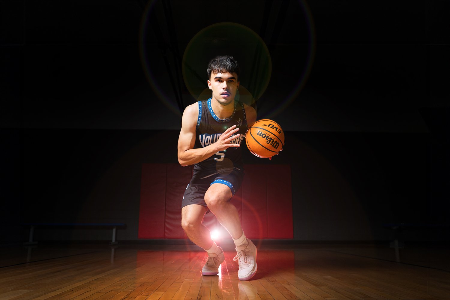 A male basketball player in a black jersey crouches with a basketball in a dark gymnasium, illuminated by a bright light from behind.