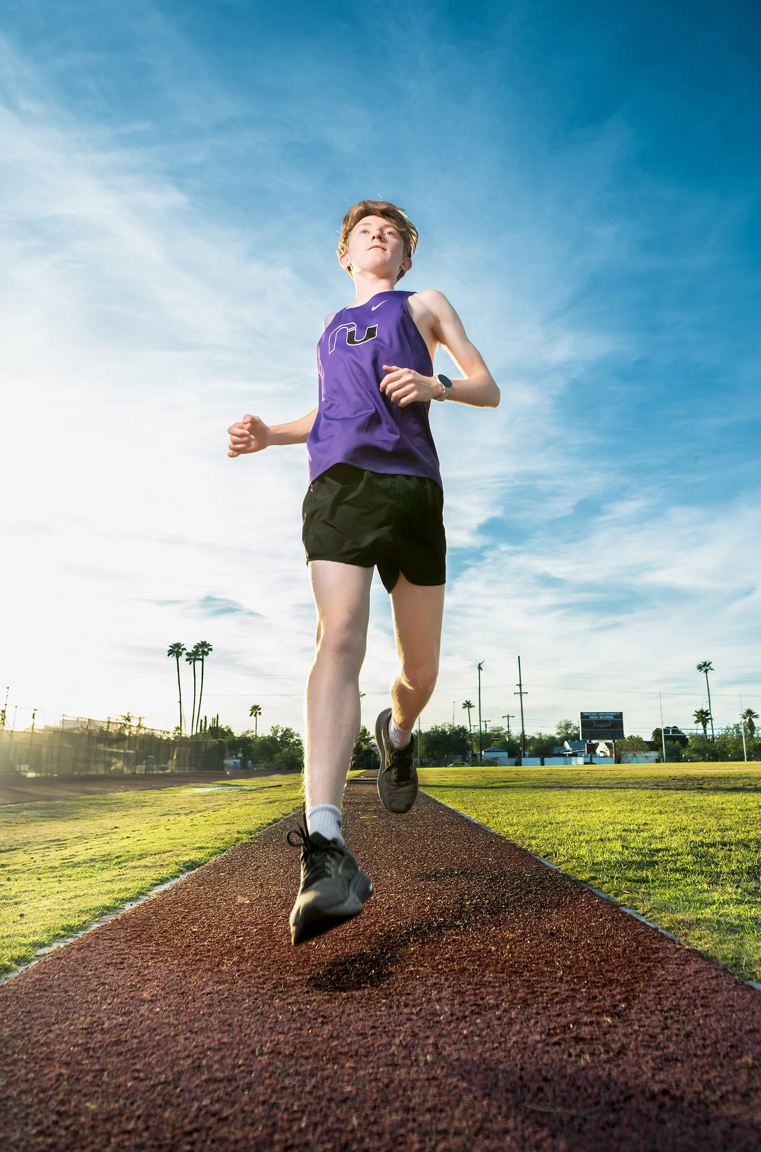 Young male runner for Rincon High in purple tank top running on a track field with palm trees and a scoreboard in the background during a late afternoon photoshoot in Tucson