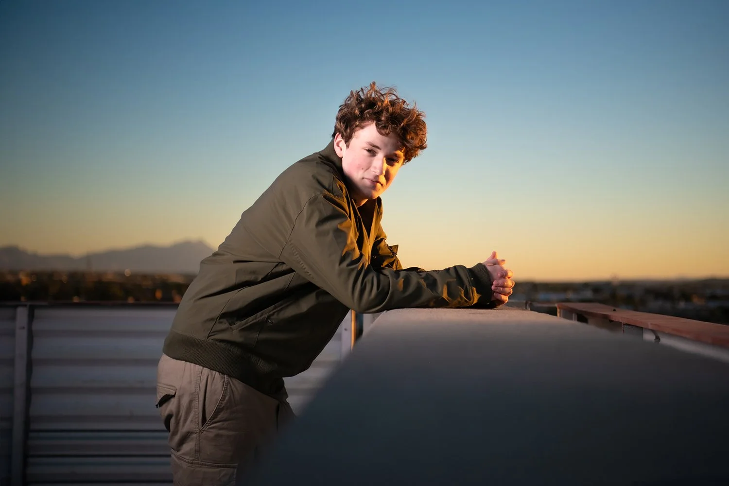 Young man with curly hair leaning on a University Arizona rooftop railing during sunset or sunrise, with mountains and cityscape in the background as part of a senior photo shoot