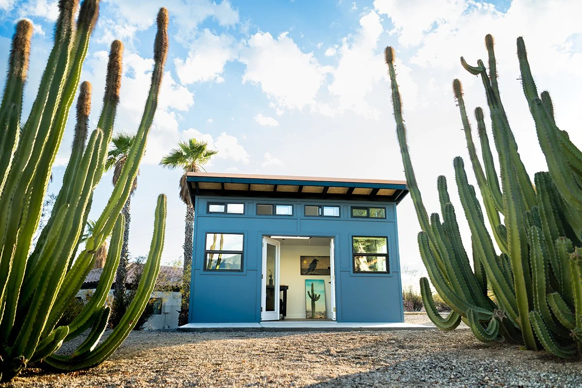 A small blue modern tiny house with a white door, surrounded by tall cacti, in a desert landscape with a palm tree and blue sky.