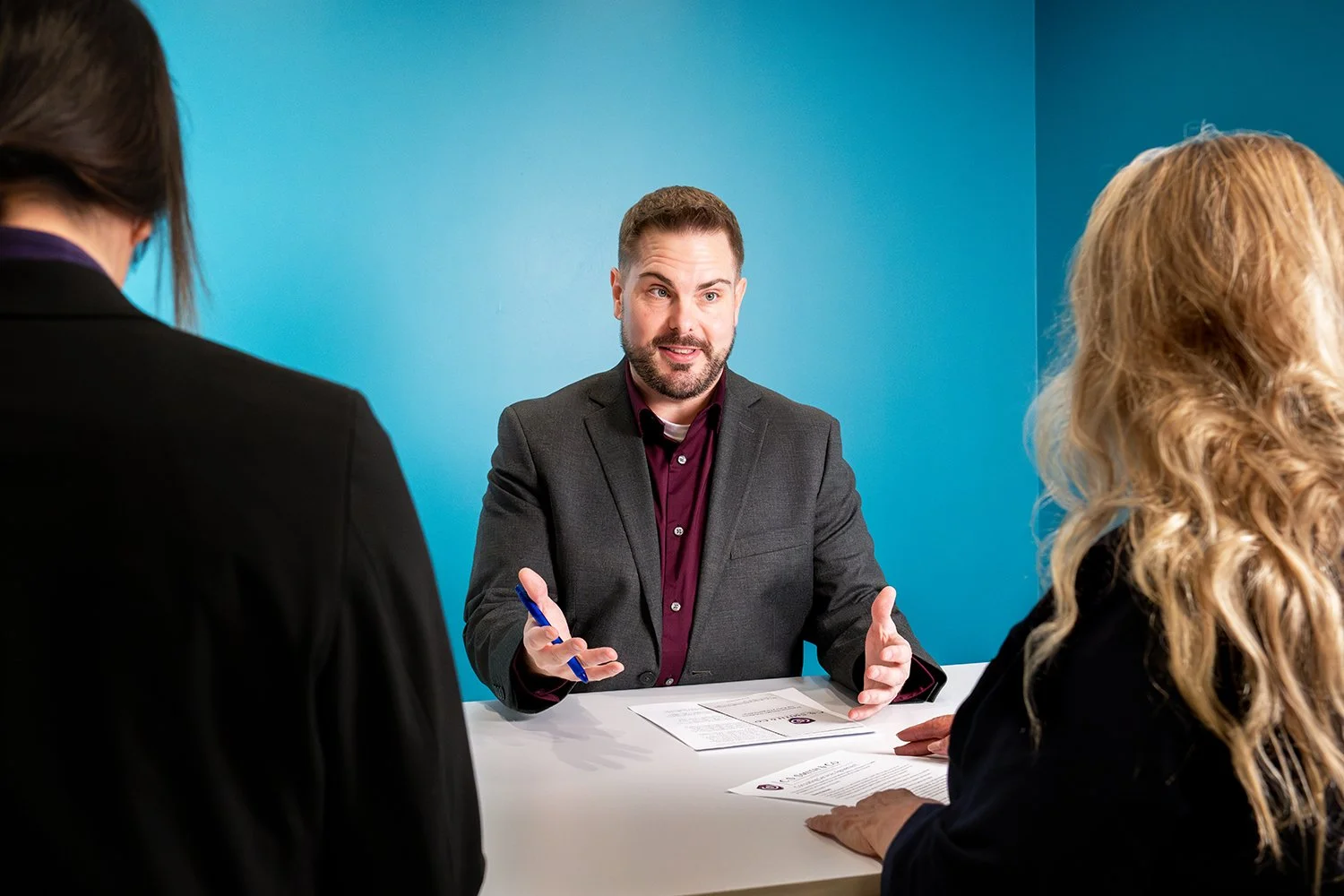 A man in a gray suit talking to two women during an interview or meeting, with a blue wall in the background.