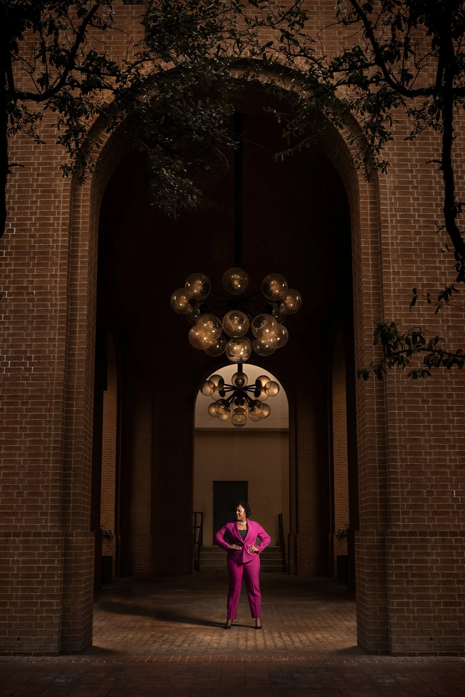 A woman in a bright pink suit stands confidently in a brick archway, with chandelier lighting above and trees partially framing the scene.