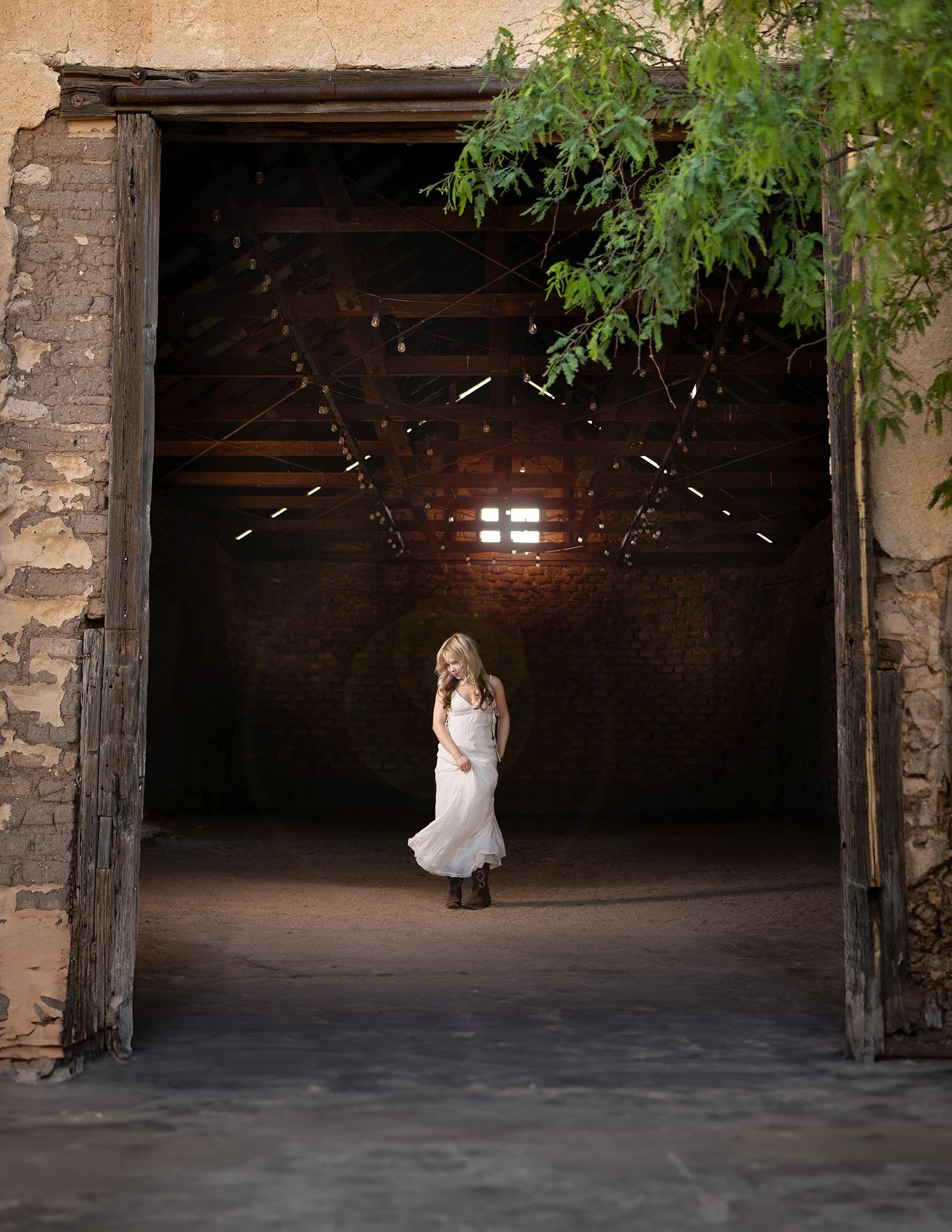 A high school senior girl in a white dress standing alone in an old barn with an open doorway, wooden beams, and string lights hanging from the ceiling, with greenery visible outside during a photo shoot in the Tucson area