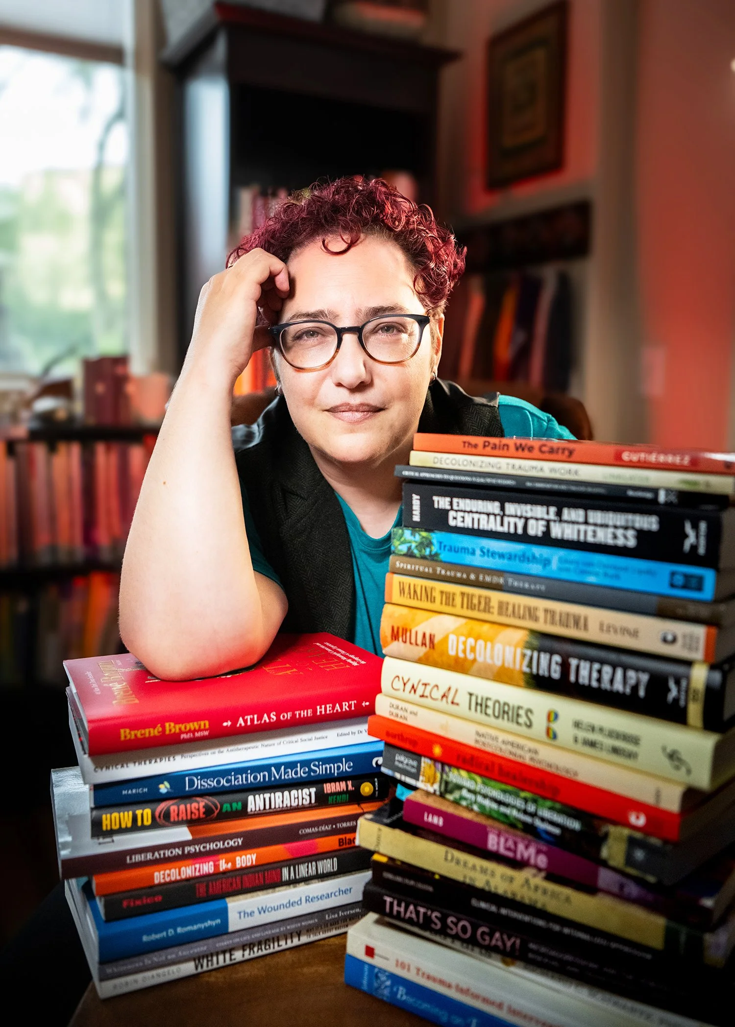 A Tucson therapist with curly red hair and glasses sits at a table with a large stack of books in front of her. The books cover topics related to psychology, social justice, decolonization, and anti-racism. The background shows a bookshelf