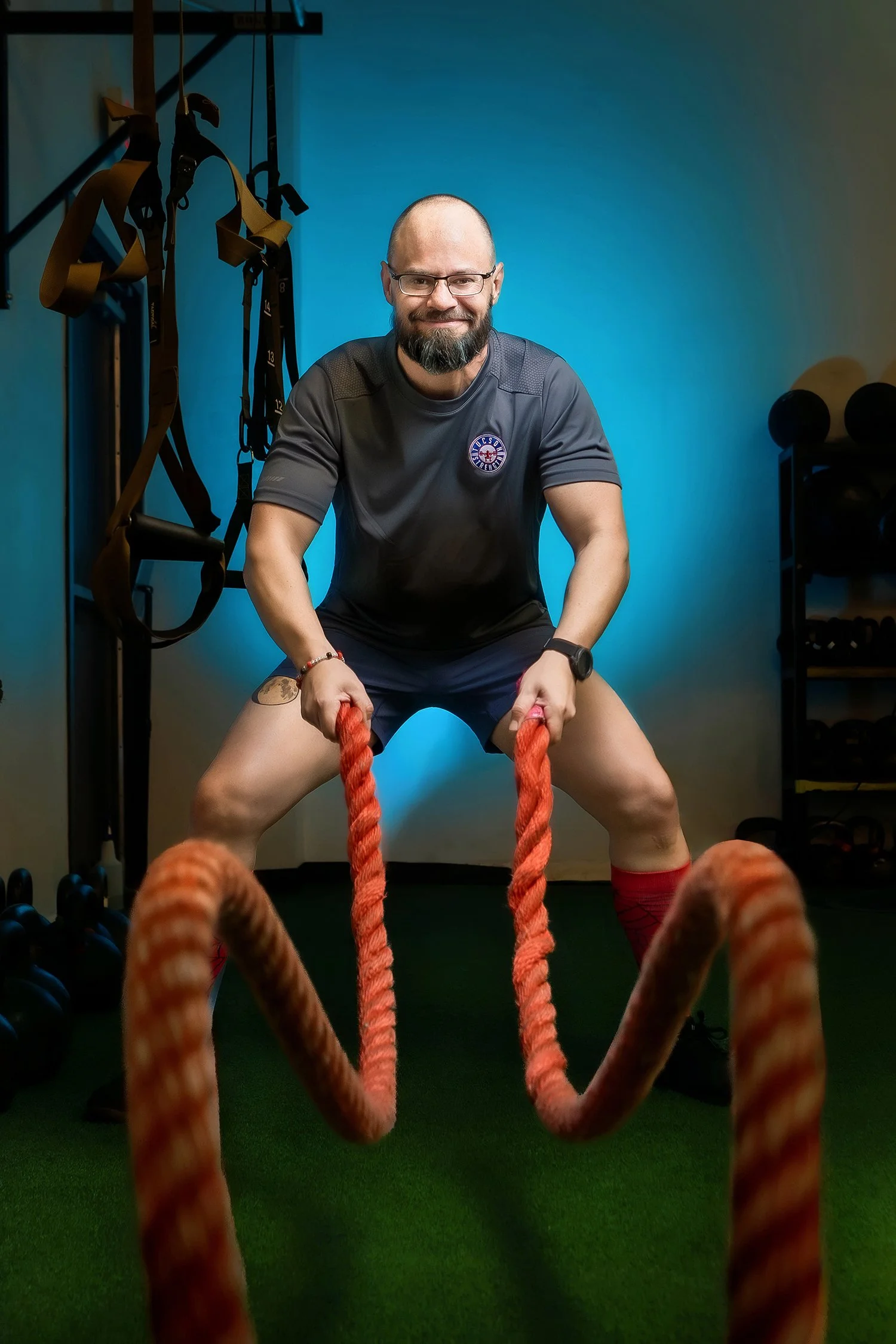 A man with a beard and glasses is working out with battle ropes in a gym. The gym has resistance bands hanging on the wall to his left, black dumbbells on a rack to his right, and a green floor. The background wall is painted blue.