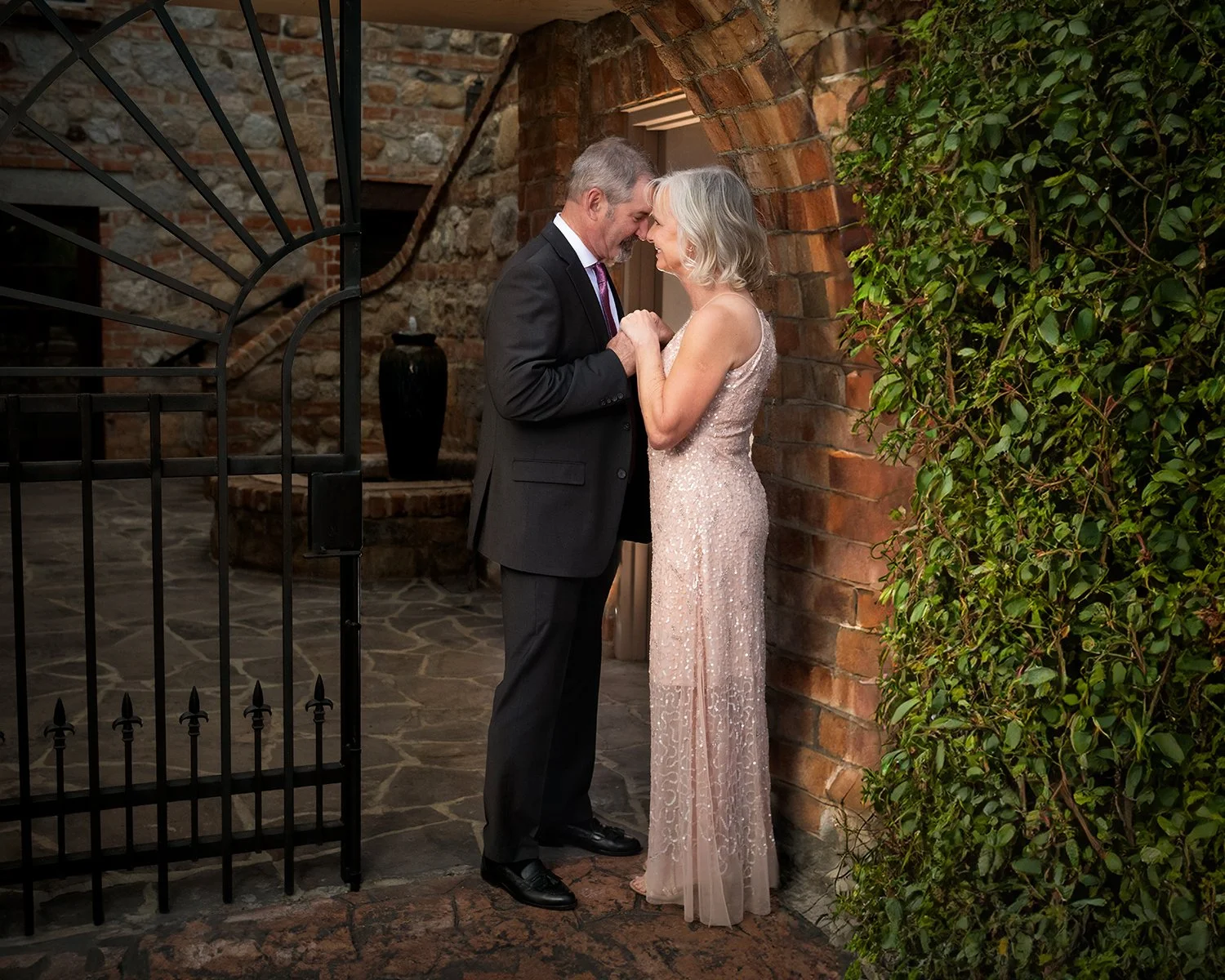 A couple dressed in formal attire sharing a moment close to a brick wall and gate, holding hands with foreheads touching at a small Tucson wedding