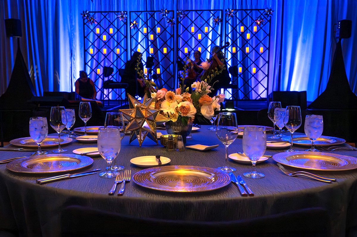 A banquet table set with gold-rimmed plates, water glasses, and silverware, decorated with a floral centerpiece and star-shaped ornaments, with a stage in the background illuminated with blue lighting and small lit candles.