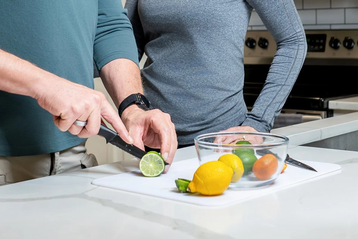 Two people preparing fresh citrus fruit, with one person slicing a lime on a cutting board and a bowl of lemons, limes, and oranges nearby.