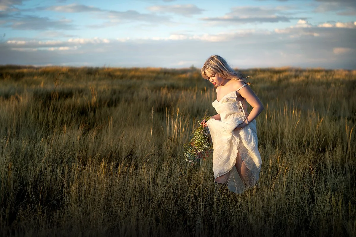 A high school senior girl in a white dress holding a small bouquet of wildflowers walking through tall grass during sunset during a tucson area senior portrait photo shoot