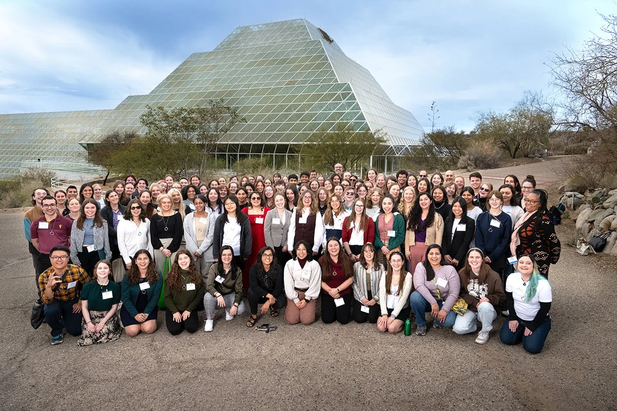 A large group of people gathered outdoors, posing for a group photo in front of a modern glass building with a pyramid-like shape, surrounded by desert landscape with sparse trees.
