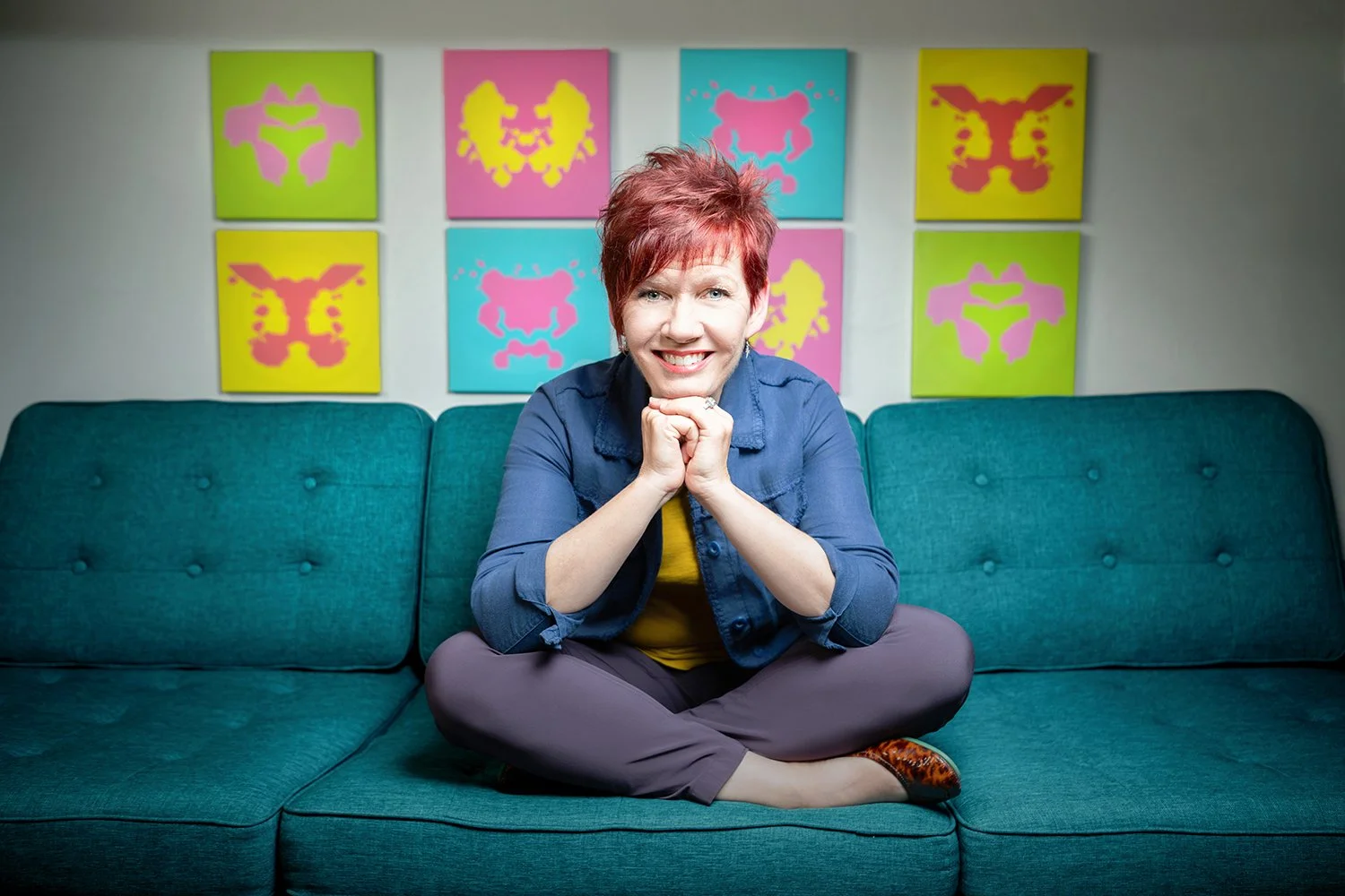 A woman with short red hair, wearing a blue jacket and gray pants, sits cross-legged on a teal sofa, smiling at the camera with hands clasped under her chin. Behind her is a wall decorated with nine colorful, abstract art pieces in bright colors like