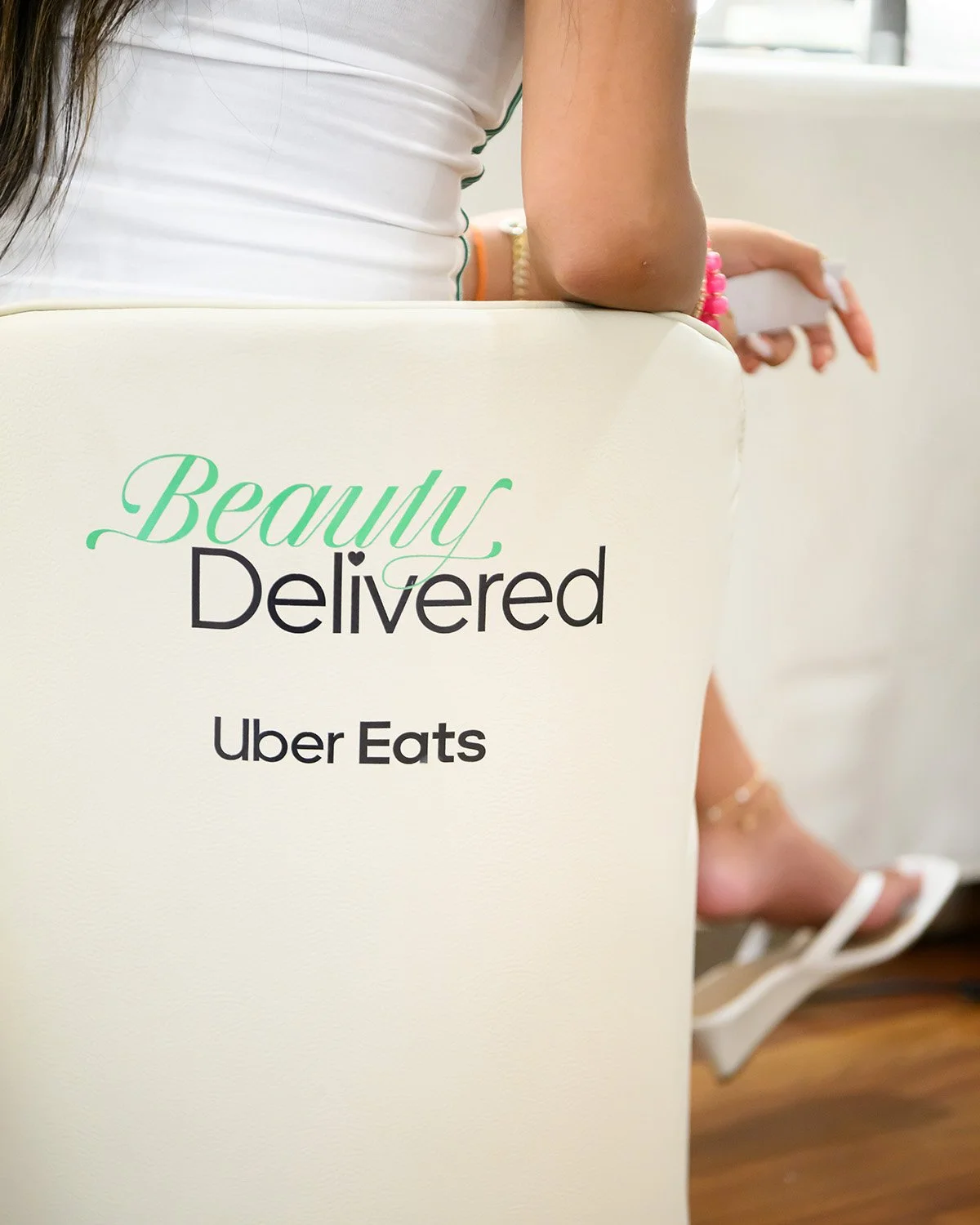 Close-up of a white cardboard box with green and black text that reads 'Beauty Delivered by Uber Eats.' A woman is sitting behind the box, holding a phone, with her arm and part of her body visible, wearing a white top, colorful bracelets, and high-h