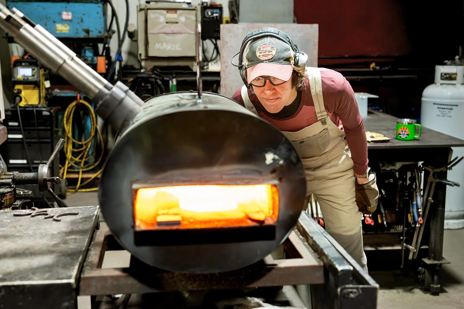 A person in safety gear observes molten metal being poured from a large industrial furnace in a workshop.
