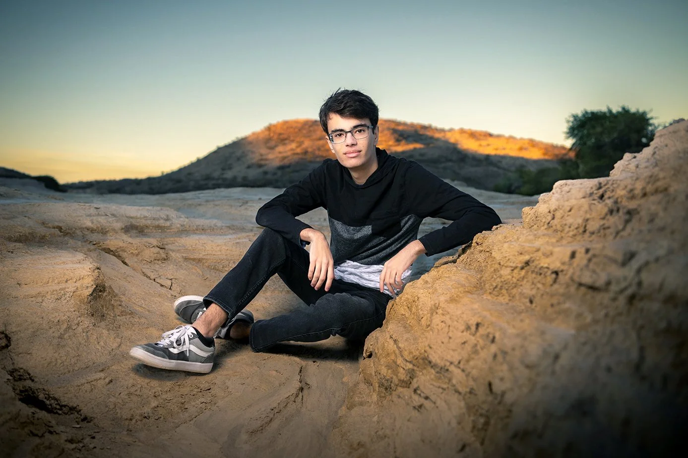Young man sitting in a rocky desert landscape during sunset, wearing glasses, a black and gray long sleeve shirt, black jeans, and gray sneakers.