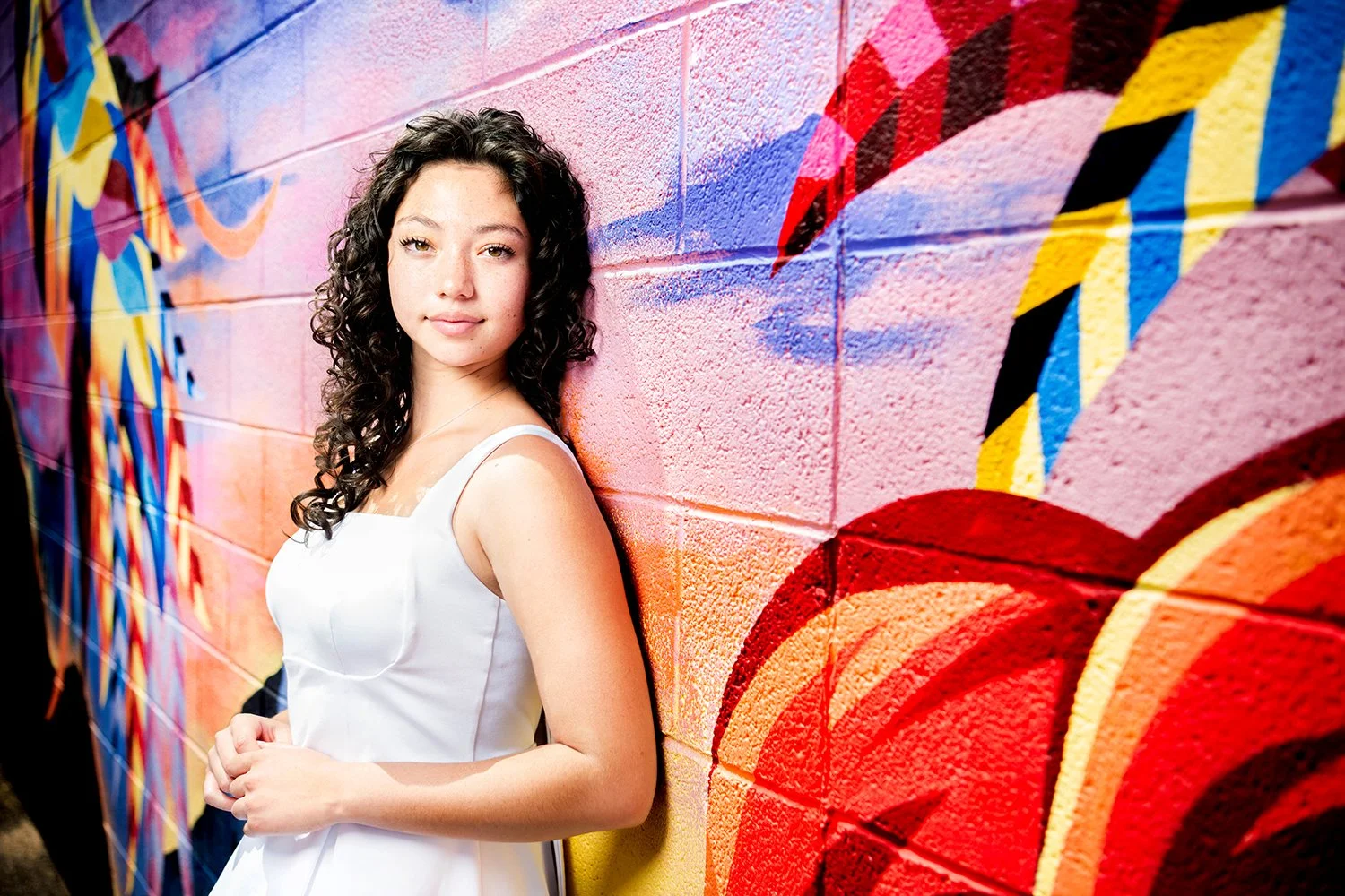 Young high school senior woman with curly dark hair in a white dress leaning against a colorful graffiti wall for her portrait photo shoot in downtown tucson, az