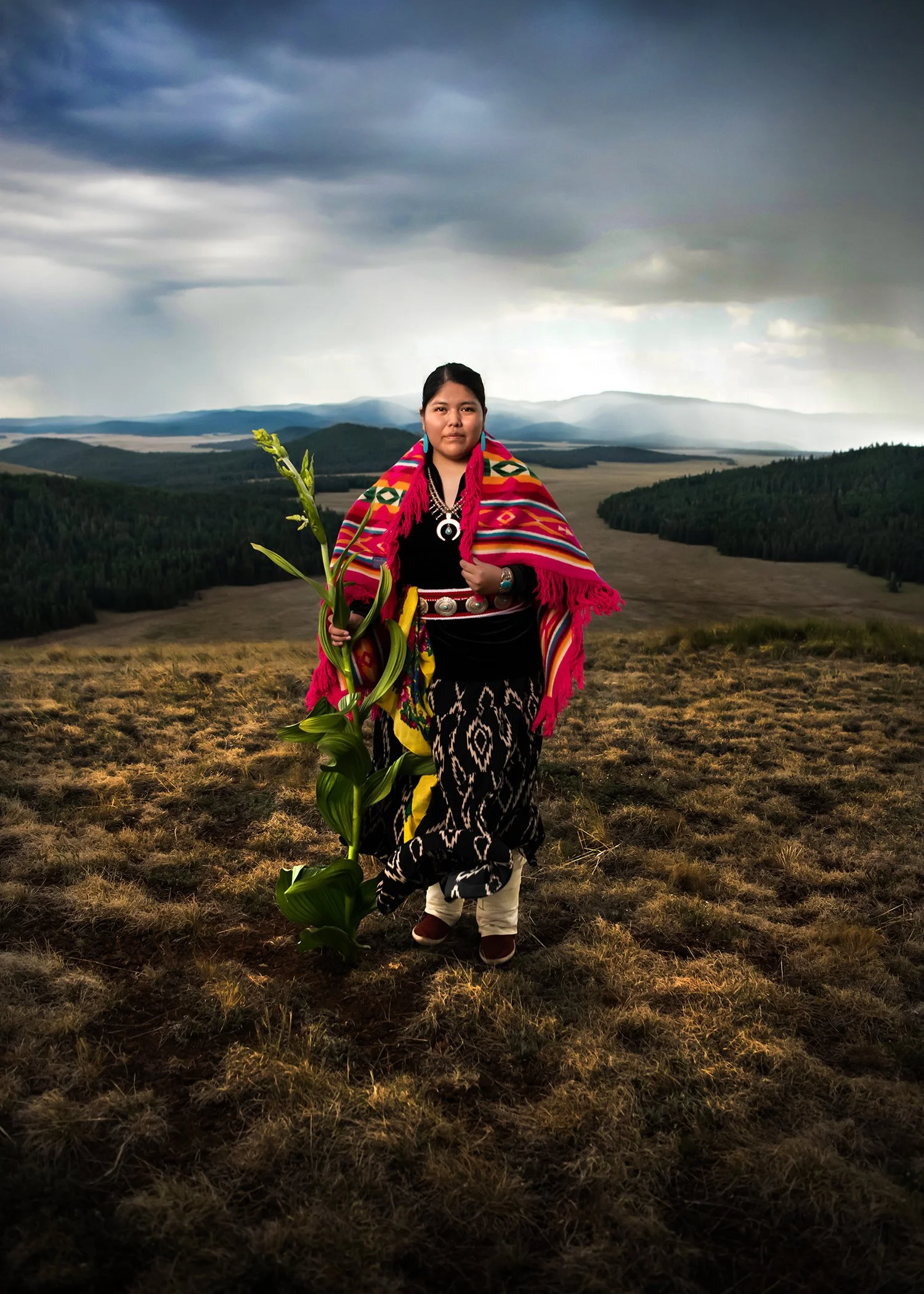A woman dressed in traditional Navajo clothing holding a green plant, standing in a field with rolling hills and dark, cloudy sky in the background for a Tucson High School senior shoot