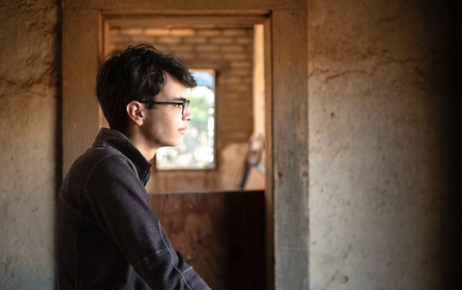 A young man with glasses, dark hair, and a black shirt sits inside a rustic wooden building, looking out a window.
