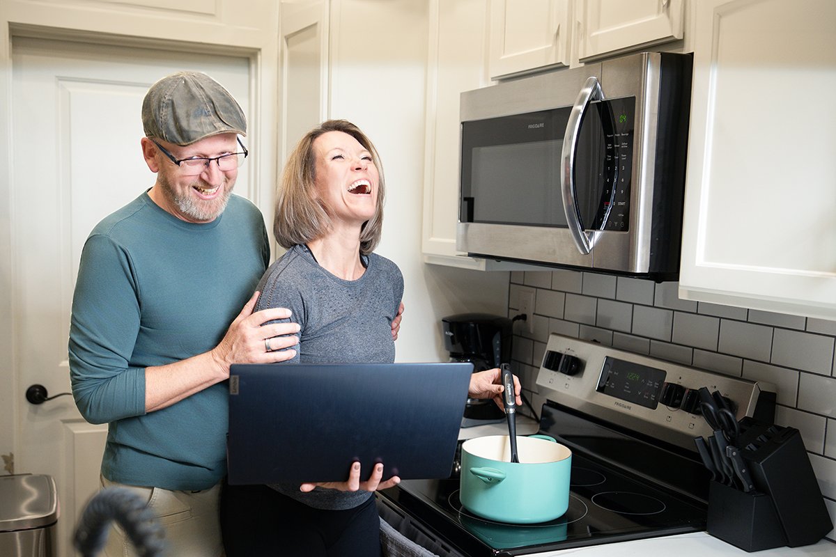 A man and woman laughing and smiling in a kitchen, with the woman holding a laptop and stirring a pot on the stove.