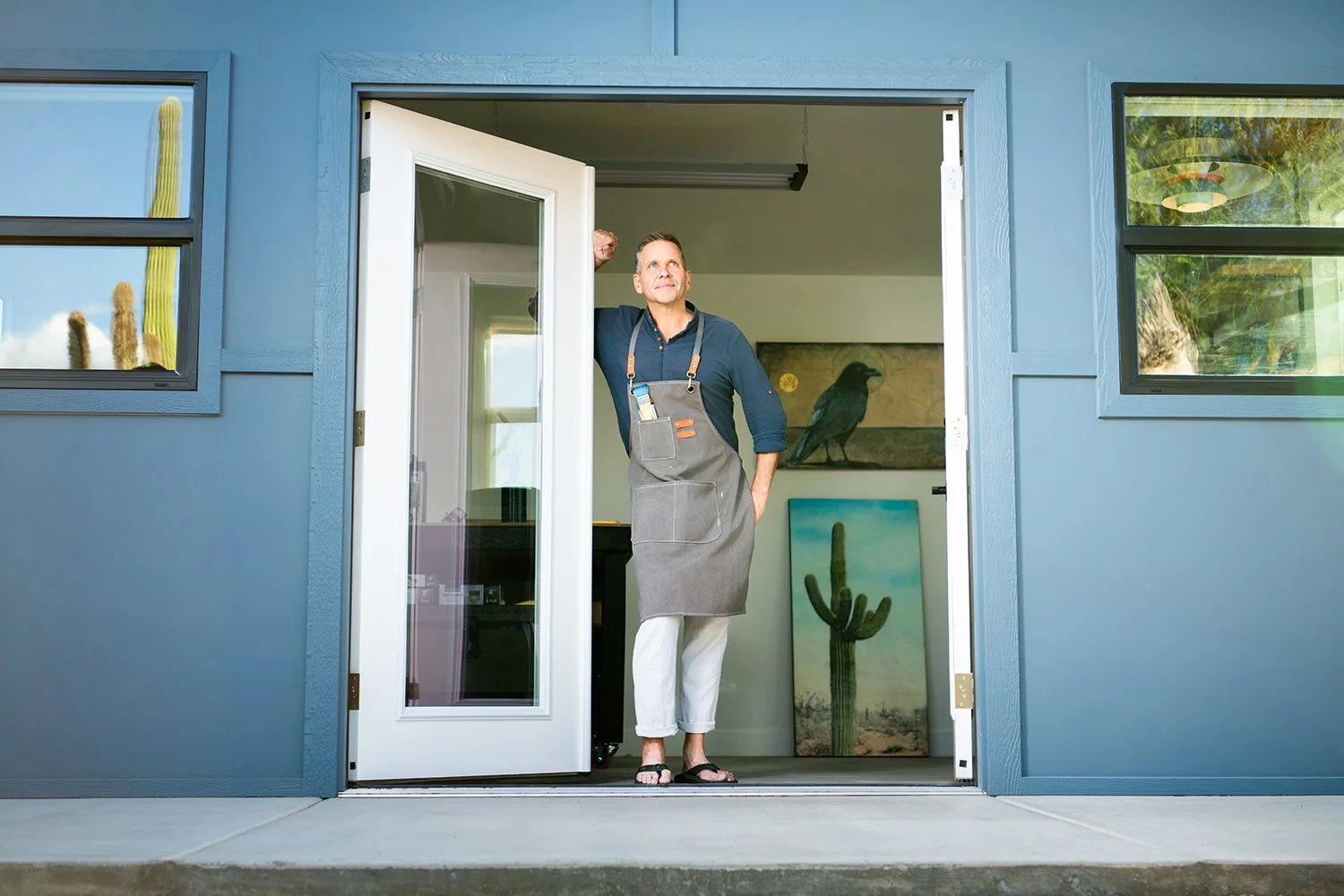 A man stands at the open door of a modern house, wearing an apron, with desert paintings and cacti visible inside.