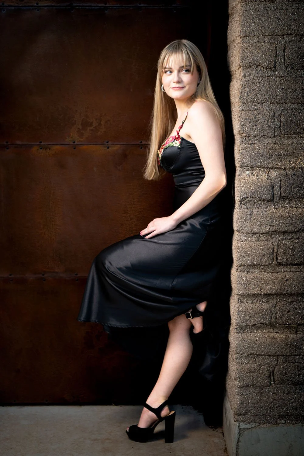 A young woman in a black dress with floral embroidery on the chest, standing against a mixed brick and wooden background, wearing black high heels and hoop earrings, with long blonde hair and a slight smile.