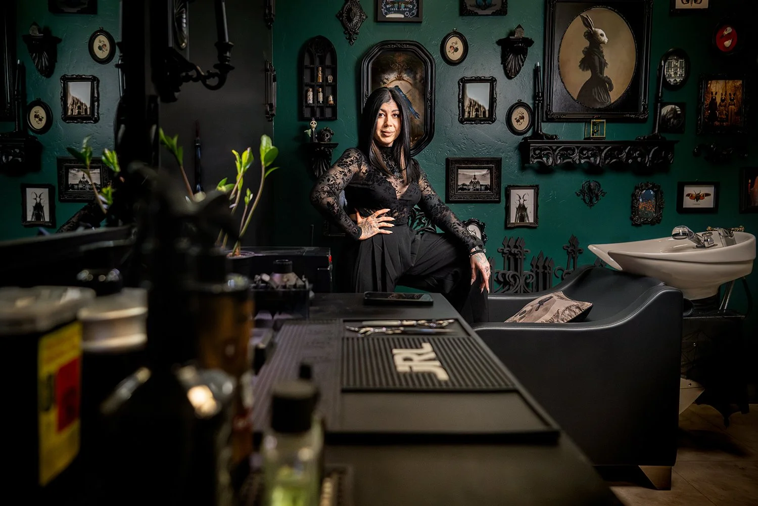 A woman with long dark hair, wearing a black lace top and black pants, posing with one hand on her hip in a room decorated with dark gothic art and black ornate frames on a green wall.