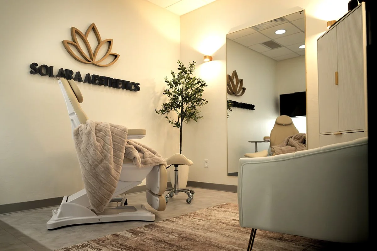 A treatment room at Solara Aesthetics with a reclining chair, a wall-mounted mirror, a potted plant, and the clinic's logo on the wall.