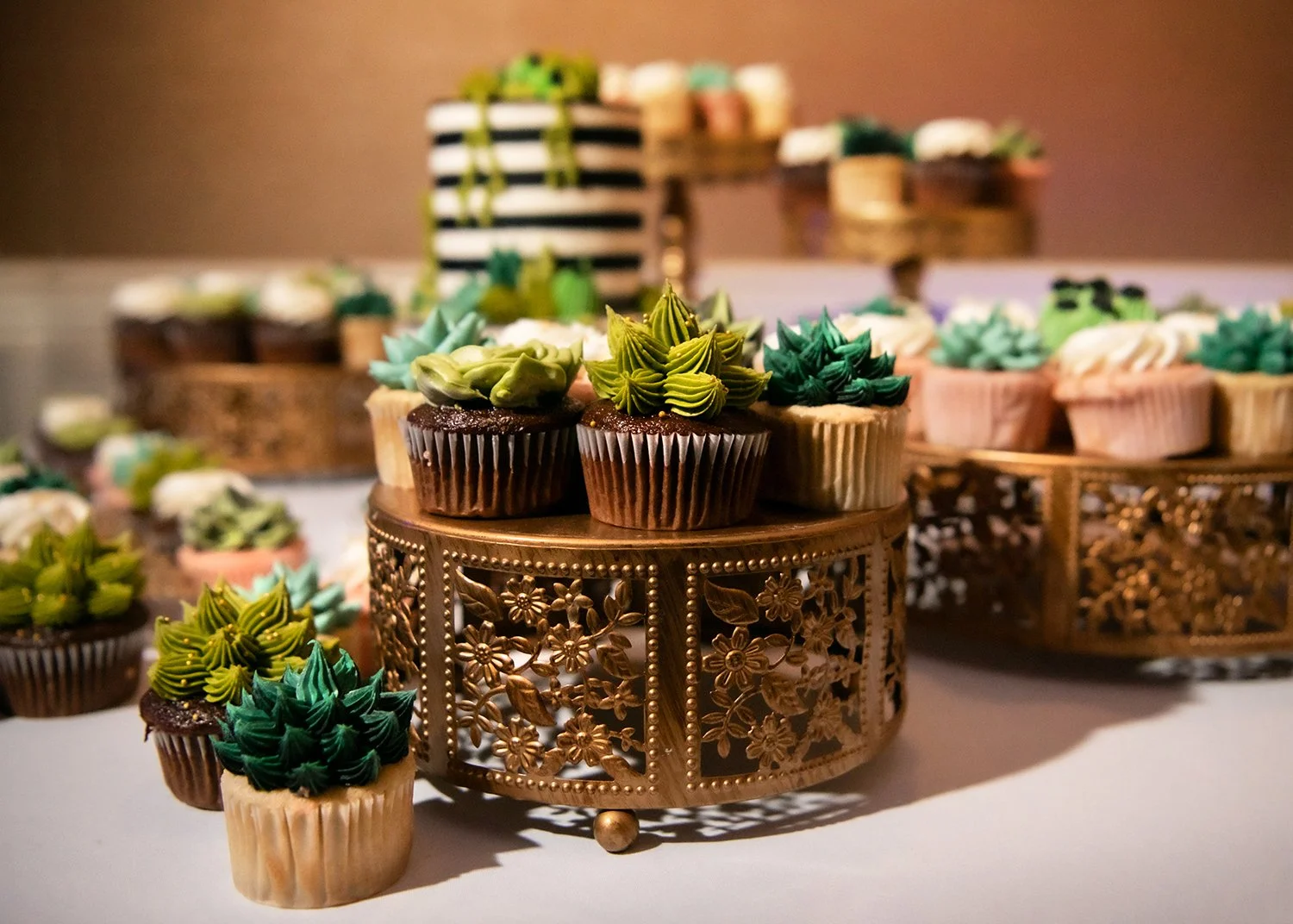 Decorative mini cupcakes with green and teal icing, displayed on ornate gold stands and wooden trays.