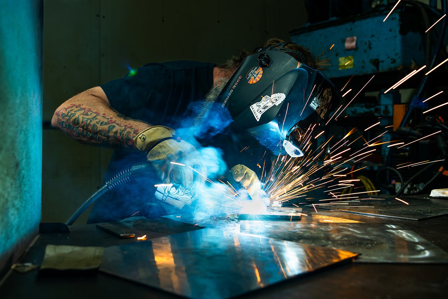 Worker welding metal on a workbench with sparks flying, wearing a welding helmet, gloves, and a black shirt, in a workshop.