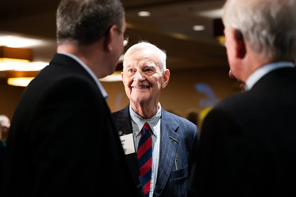 An elderly man with white hair smiling and talking with two other men at a formal event.