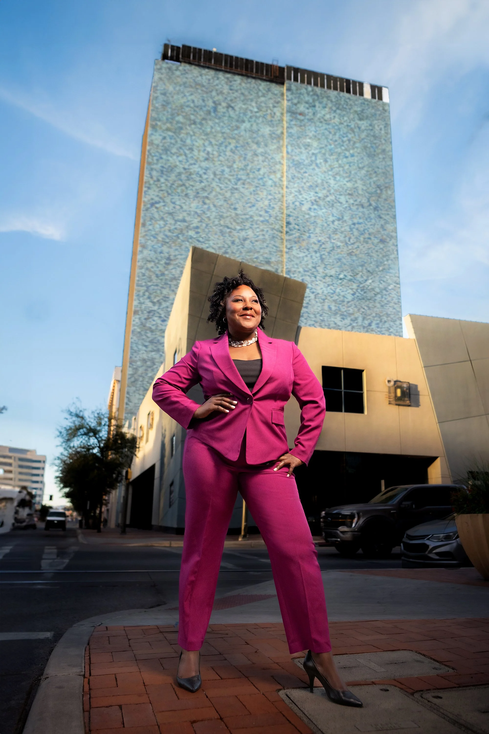 A woman with short curly hair stands confidently on a city street corner, wearing a bright pink pantsuit and black high heels, with tall buildings in the background.