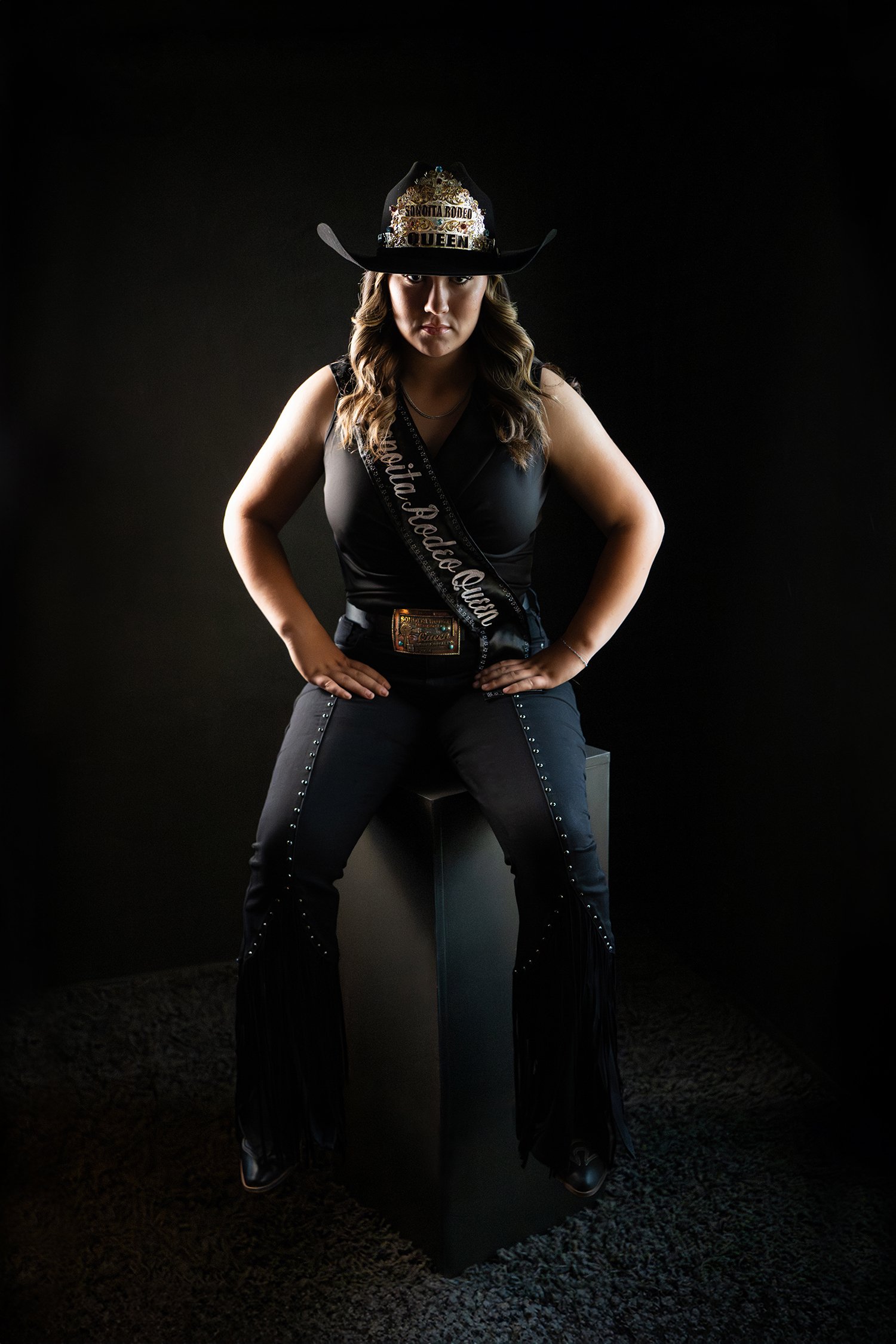 Woman wearing a black cowboy hat, black sleeveless top, and black pants with silver studs, standing with hands on hips against a dark background. The hat has the words 'Sonoita Rodeo Queen' during a Tucson senior portrait photo shoot 