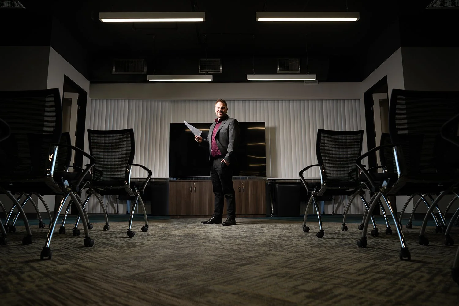 A man in a business suit standing in a conference room, holding papers and smiling, surrounded by empty chairs.