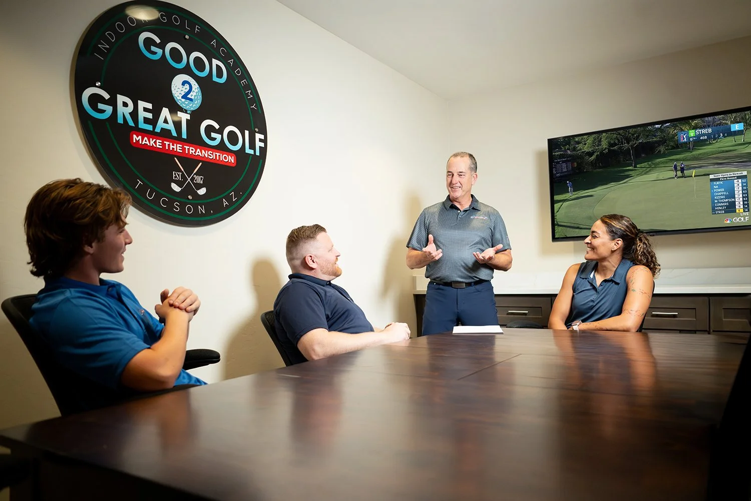 A group of four people in a meeting room, with two women and two men, sitting around a dark wooden table. One man is standing and speaking, while the others are listening and smiling. There is a large TV screen on the wall showing a golf game, and a 