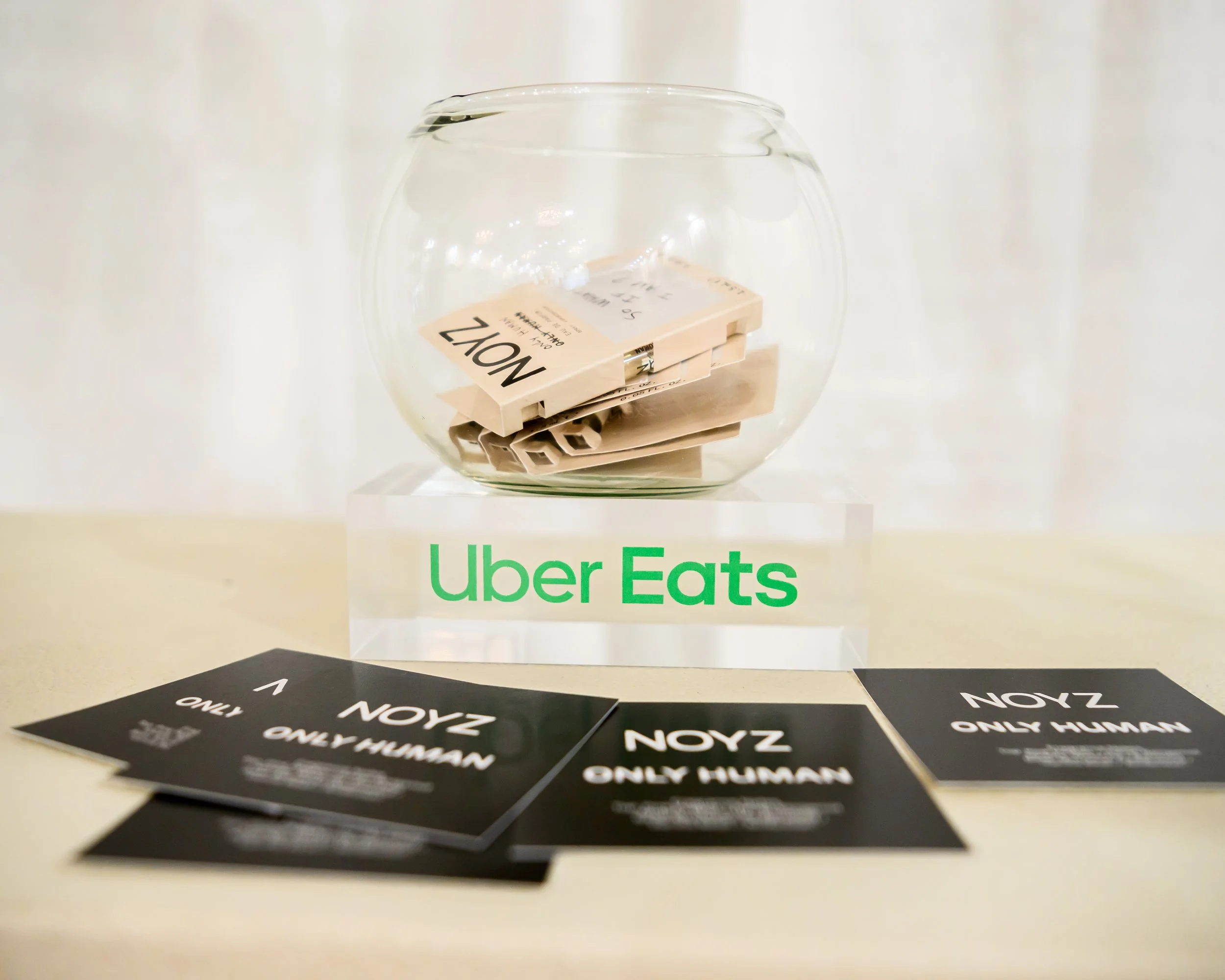 A clear glass fishbowl containing folded paper notes with one visible marked 'NOZ' and some notes beneath, placed on a surface with several black stickers reading 'ONLY HUMAN' scattered around.