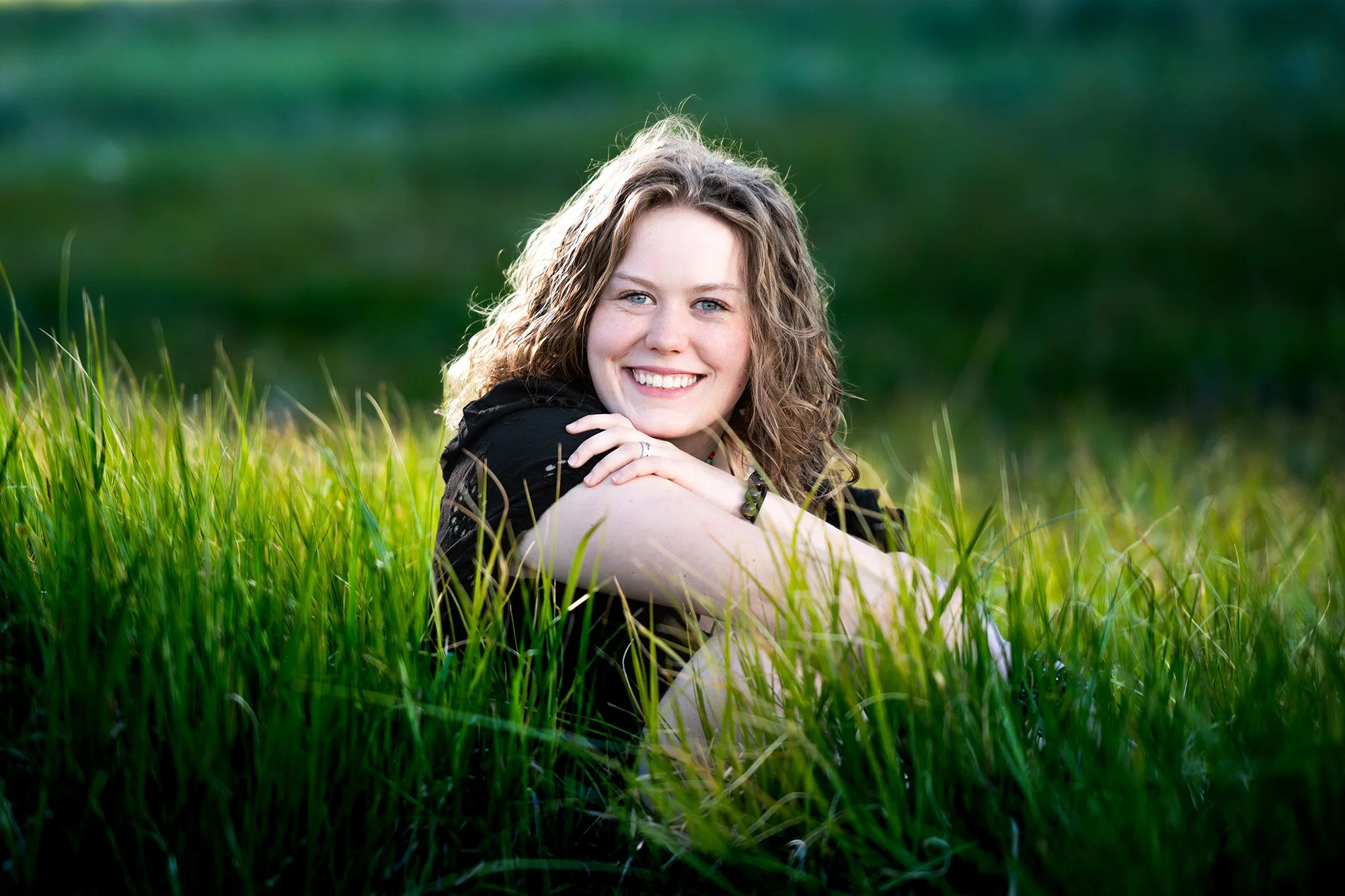 A woman with curly blonde hair smiling and resting her chin on her hand in a grassy field with a green landscape background during a Tucson high school senior photo shoot