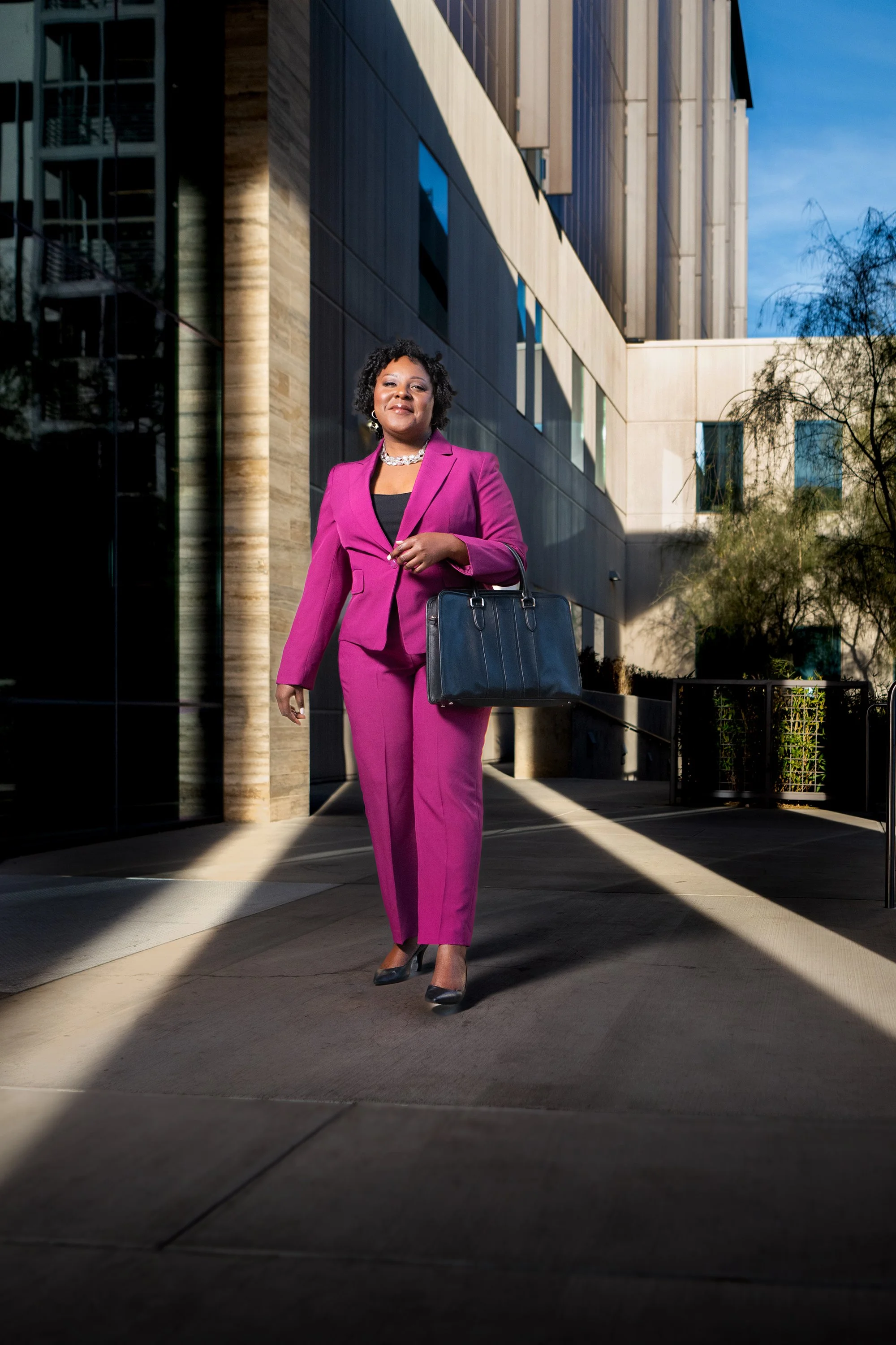 A confident woman in a bright pink suit walking outdoors near a modern building during daytime, carrying a black handbag.