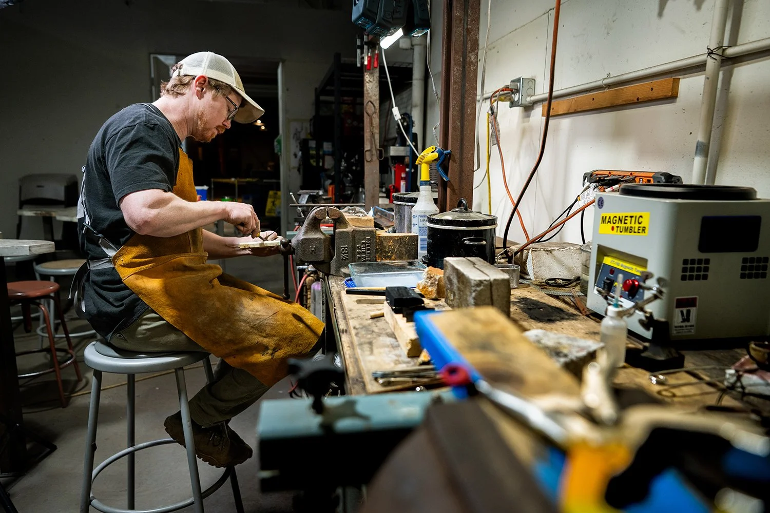 A man wearing glasses, a baseball cap, and a brown apron is working at a workbench in a workshop, surrounded by tools, equipment, and materials.