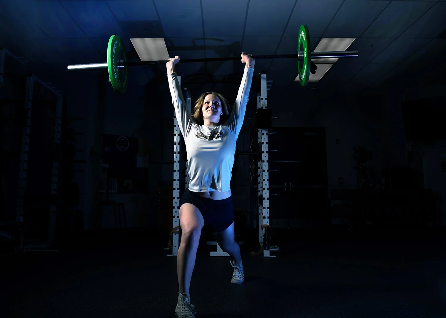 A young woman lifting a barbell overhead in a high school gym, with a focused expression, dark background, and gym equipment visible as part of her senior photo shoot in Tucson