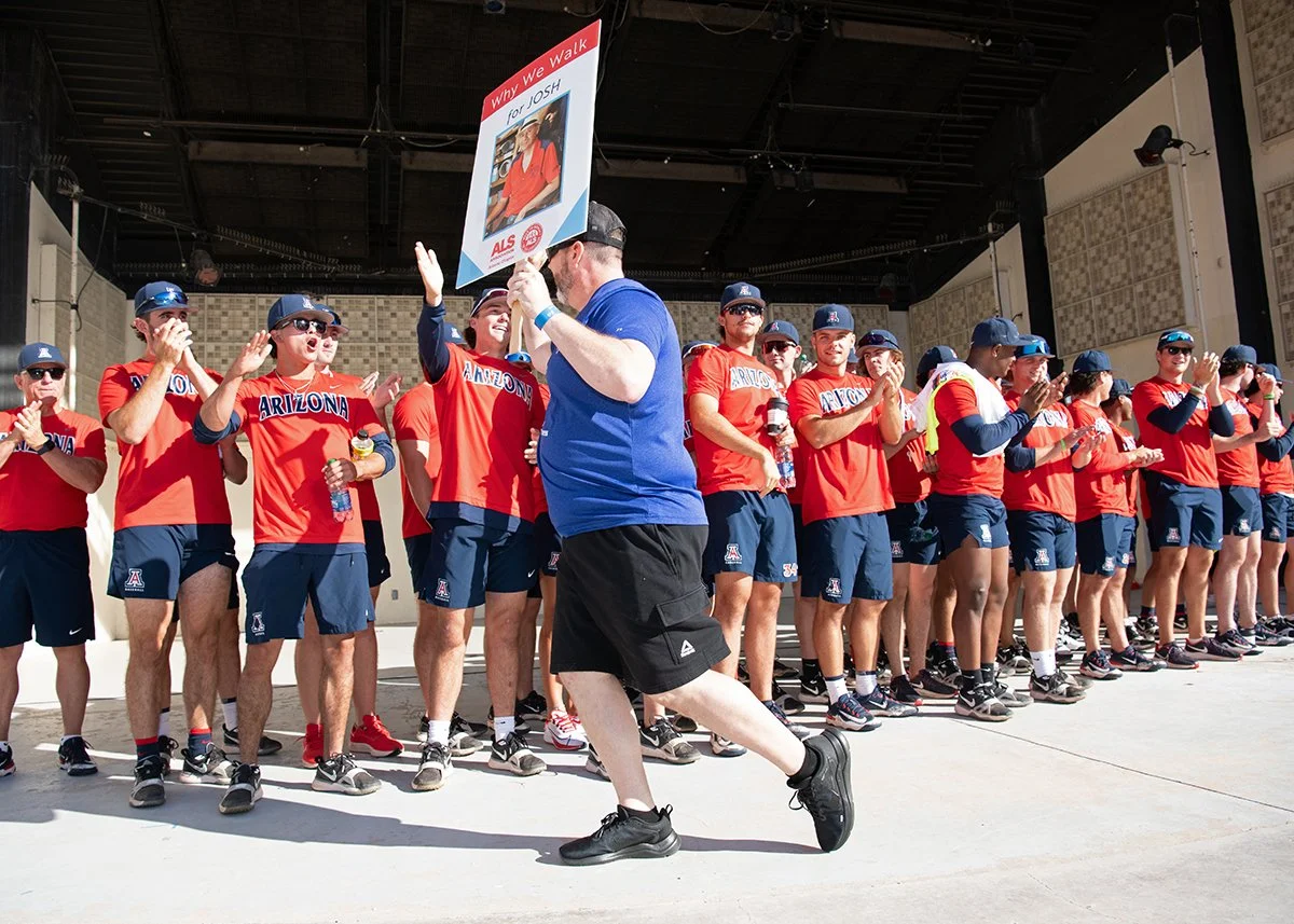 Arizona softball team in red shirts and blue shorts standing in a line, clapping and celebrating, as a man in a blue shirt and black shorts walks past holding a sign that says 'Why We Walk for JOSH' with a photo of a man.