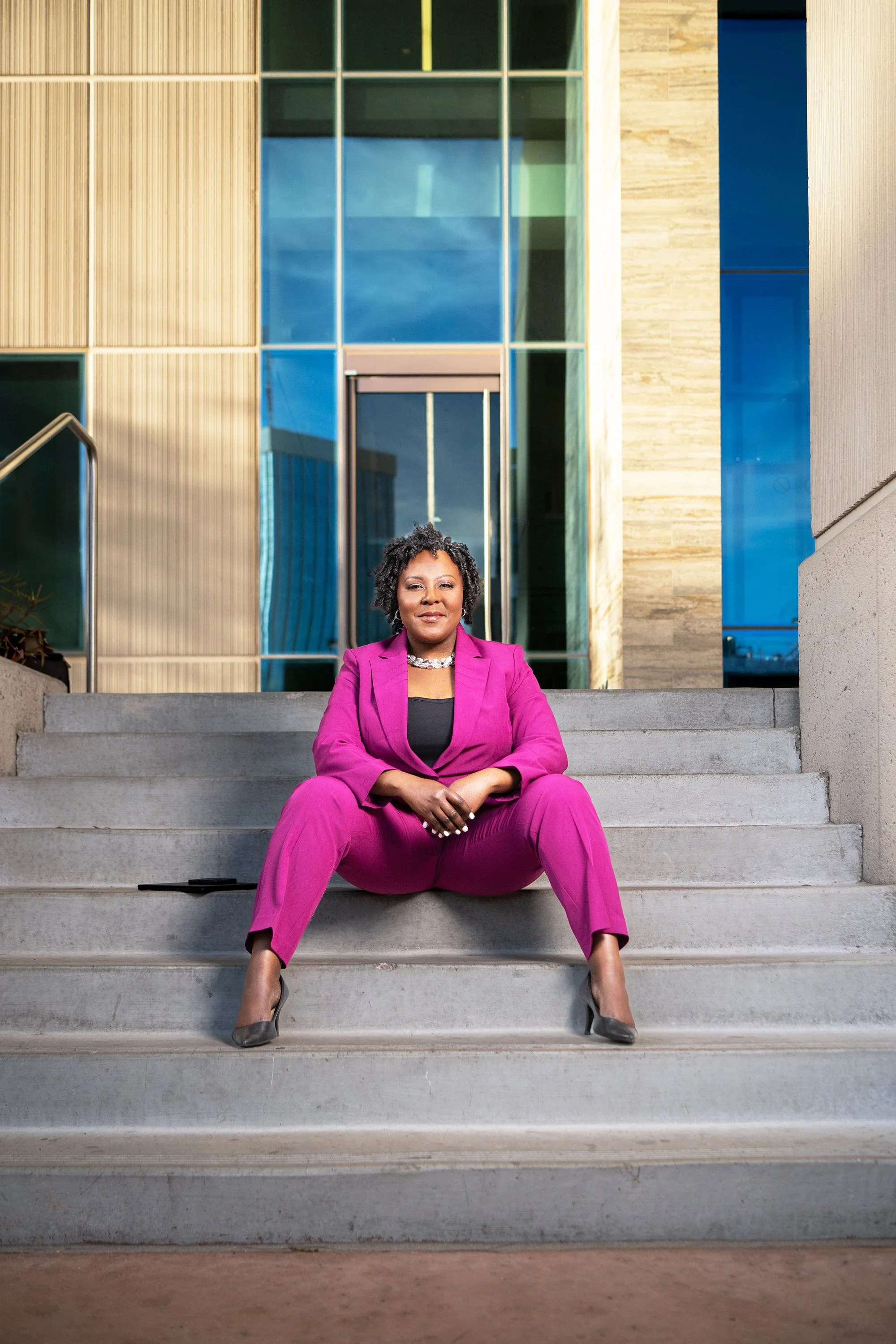 A woman with curly black hair in a pink suit sitting on stairs in front of a modern building with large glass windows and wooden panels.