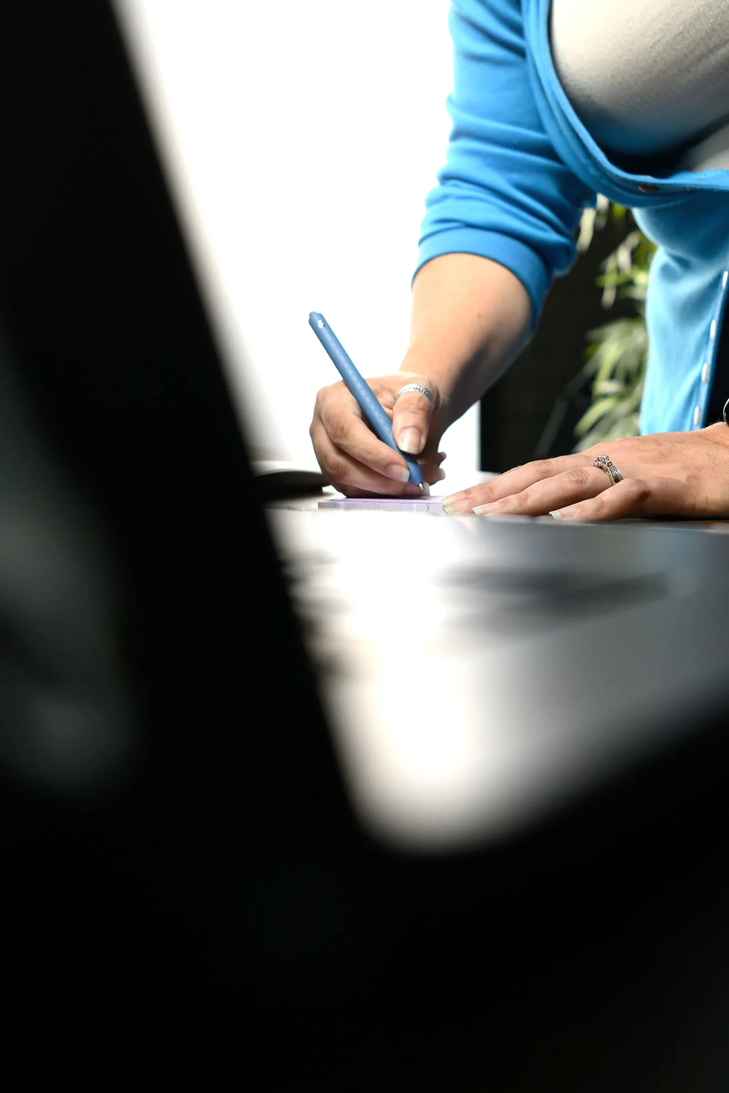 A person wearing a blue jacket is signing a document with a blue pen on a table. The focus is on their hand and the pen, with focus on the jewelry including a ring and a bracelet. The background is blurred, with some green foliage visible.